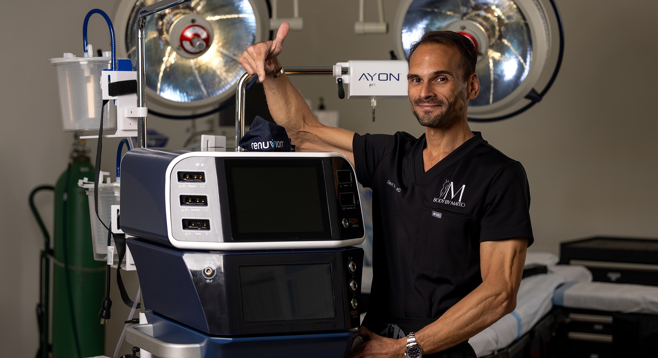 Man in scrubs beside medical equipment, giving thumbs up.